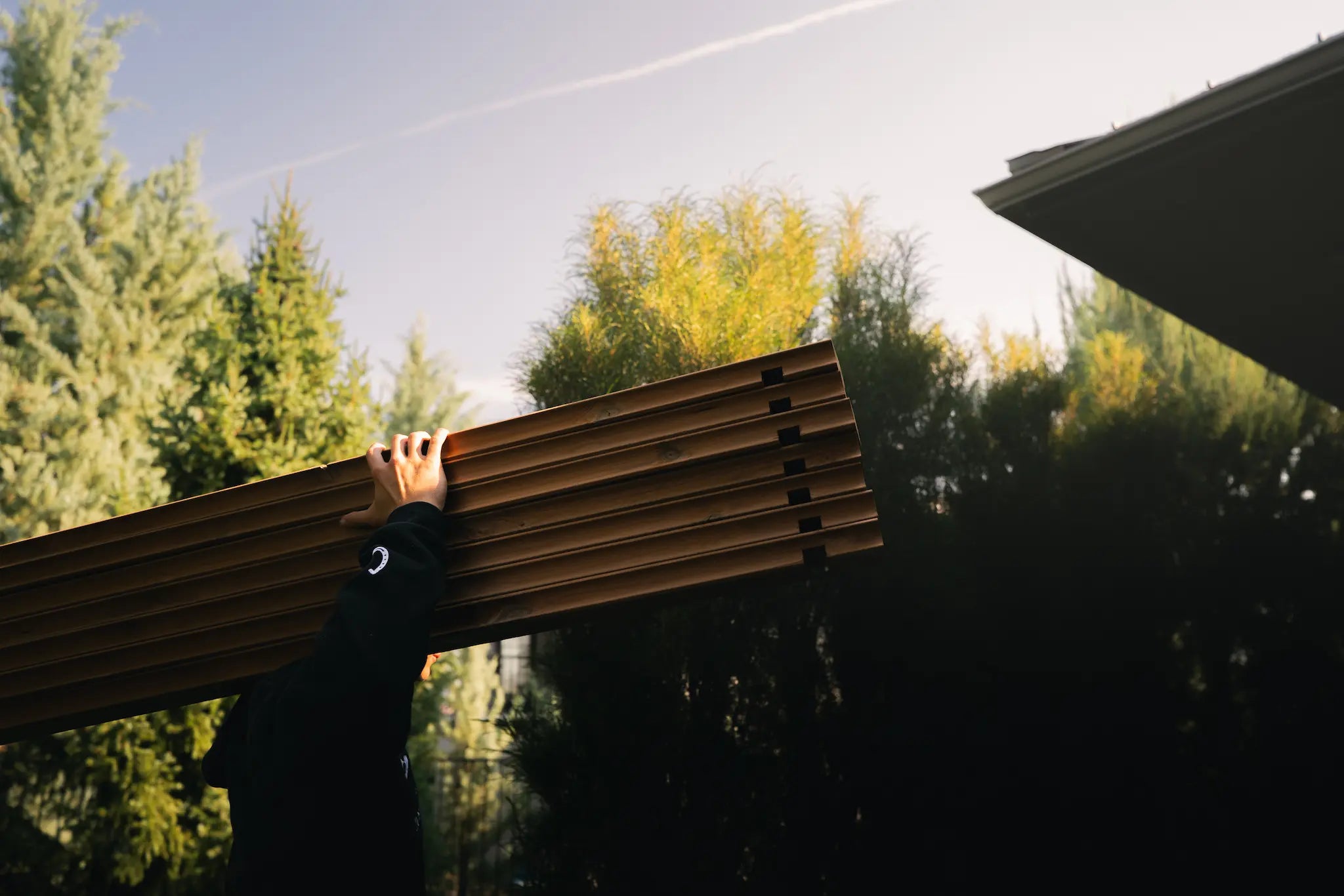 Person carrying wooden slats outdoors with trees and a building in the background.